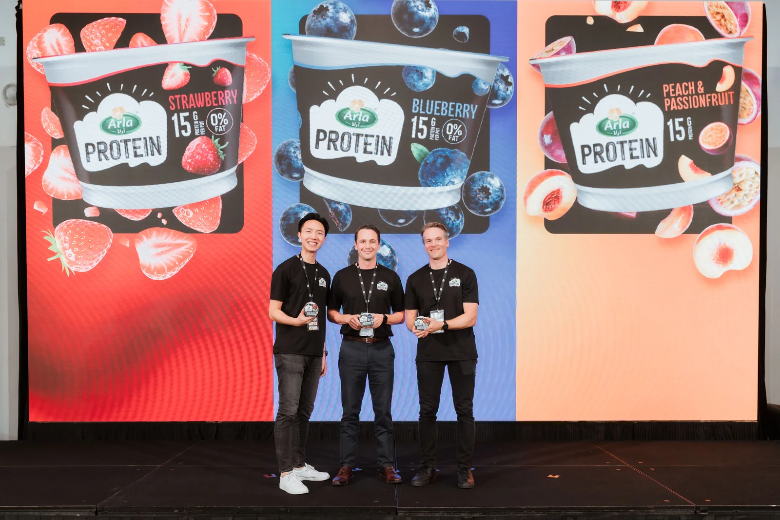 Three Arla presenters holding the strawberry, blueberry, and peach and passionfruit Arla Protein cups on stage