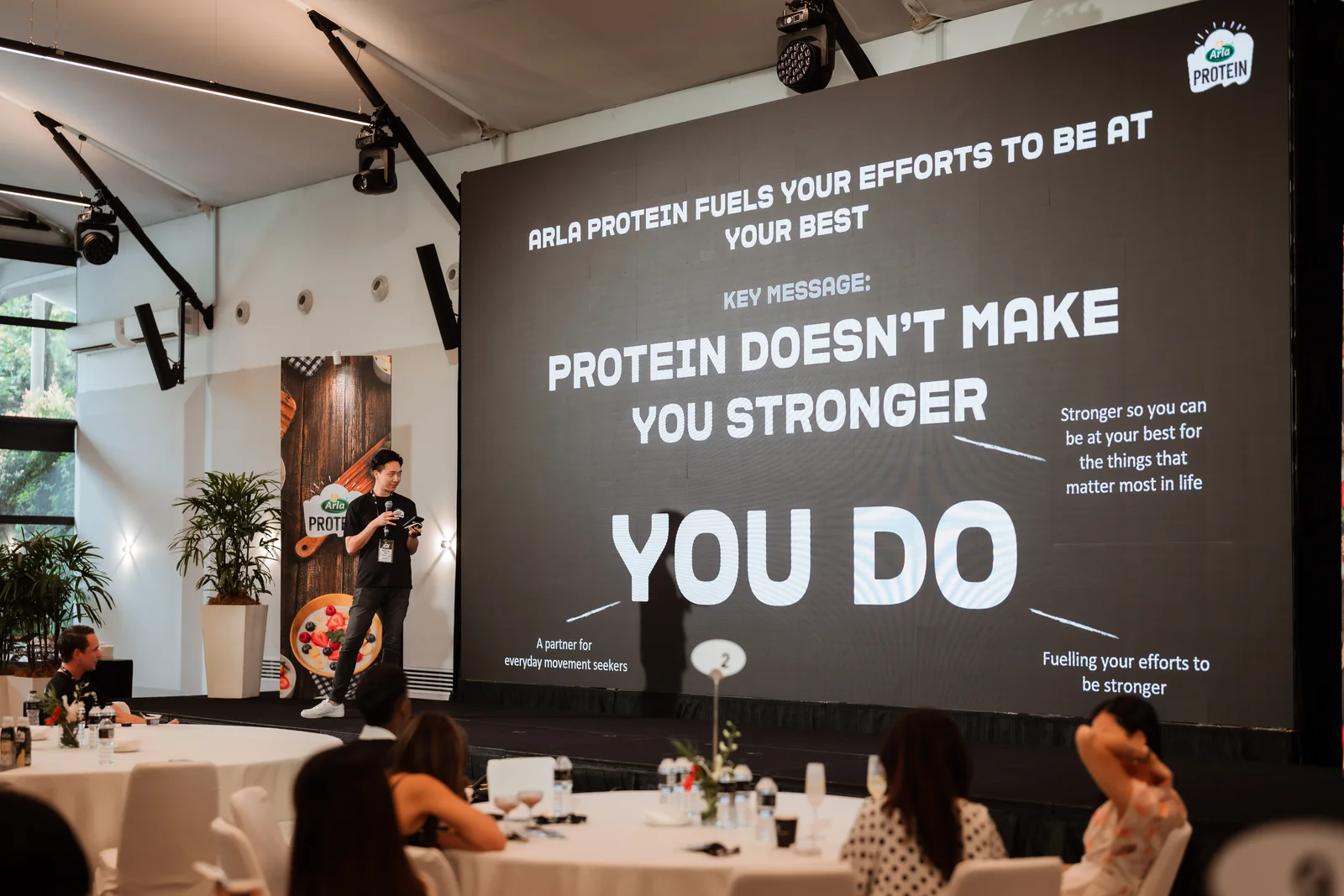 A presenter on stage in front of the Arla Protein campaign line, Protein doesn't make you stronger. You do.
